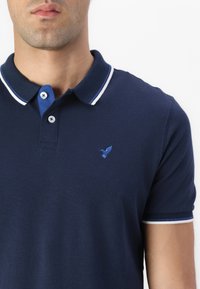 Navy polo shirt made from cotton, featuring a collar with white and blue accents, two buttons, and a small blue bird logo on the chest.