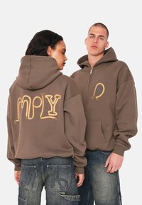 Brauner Hoodie mit geprägtem, seilähnlichem goldenen Schriftzug auf dem Rücken und einem Reißverschluss vorne, kombiniert mit blauen Jeans.