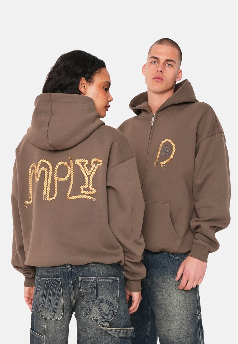 Brauner Hoodie mit geprägtem, seilähnlichem goldenen Schriftzug auf dem Rücken und einem Reißverschluss vorne, kombiniert mit blauen Jeans.