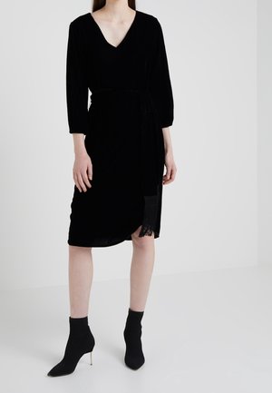 Kobieta ubrana w czarną sukienkę sięgającą kolan z rękawami do połowy przedramienia, dekoltem w serek, wiązaną w talii oraz czarne, spiczaste botki na wysokim obcasie.