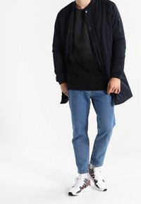 Giubbotto bomber nero con un maglione nero a trama sotto, jeans blu e sneakers bianche con dettagli rossi e neri.