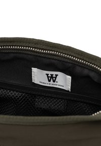 Pungă cu fermoar de culoare verde măsliniu, cu interior negru, dotată cu un buzunar din plasă și un eticheta albă cu text negru care indică "DOUBLE V BY WOOD WOOD."