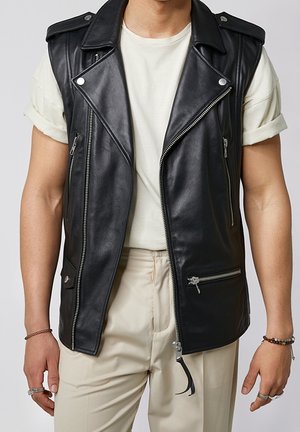 Mężczyzna w czarnej skórzanej kamizelce bez rękawów na białym t-shircie z podwiniętymi rękawami i beżowych spodniach, z bransoletkami i pierścieniami na rękach.