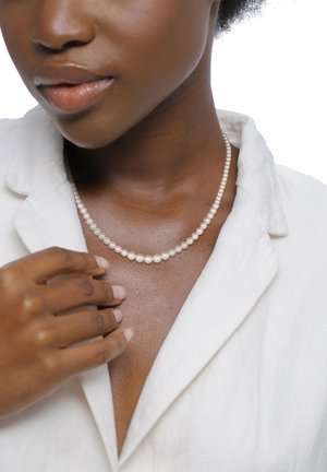 Collier de perles court avec des perles blanches rondes, porté sur une peau bronzée. La personne porte un haut blanc à col ouvert, avec la main reposant doucement sur le cou.