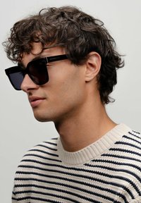 Černé sluneční brýle ve čtvercovém tvaru s tmavými skly, s elegantním designem. Modelka má na sobě svetr v krémovém a námořnickém pruhu.