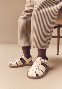 Sandales en cuir blanc avec broderie florale, design à bout ouvert, sangle ajustable et semelle marron. Pantalon beige à rayures complétant la chaussure.