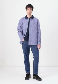Light purple cotton jacket with button closure, chest pocket, and logo patch. Worn over a dark shirt and paired with blue jeans and black shoes.
