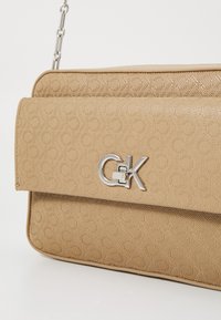 Beige käsilaukku tekstuurilla ja hopeisella logo-yksityiskohdalla. Siinä on läppäkiinnitys ja irrotettava ketjuhihna.