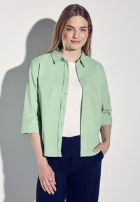 Ljusgrön skjorta med knappar och uppkavlade ärmar i trekvartslängd, bärs över en vit topp, i kombination med marinblå byxor. Mjuk textur och avslappnad design.