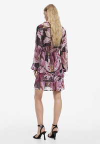 Durchsichtiges Kleid mit floralem Muster in verschiedenen Rosatönen, mit langen Ärmeln, tailliertem Schnitt und ausgestellt Rocksaum. Getragen mit schwarzen Riemchenschuhen.