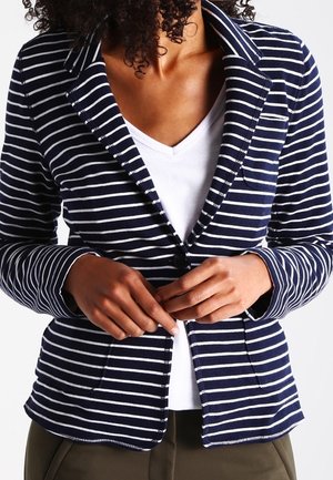 Mujer con una chaqueta azul marino y blanca de rayas sobre una camisa blanca con cuello en V abrochándose el botón de la chaqueta con ambas manos.