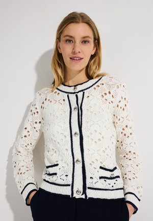 Femme aux cheveux blonds portant un cardigan en crochet blanc avec des bordures noires et des boutons argentés, les mains dans les poches, fond neutre.