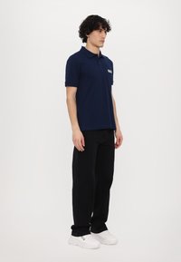 Mornarska polo majica iz bombaža s kratkim ovratnikom in logotipom na levi strani prsnega dela, v kombinaciji s črnimi hlačami in belimi supergami.