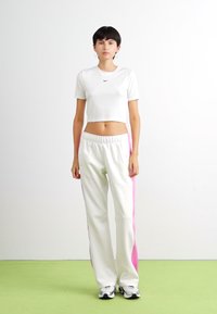 Balts īsās piedurknes t-krekls ar melnu Nike logo, savienots ar baltām sporta biksēm, kurām ir rozā sānu svītras. Gluda auduma, brīvs piegriezums.