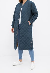 Personne portant un long manteau matelassé bleu marine avec des boutons-pression, par-dessus un haut blanc, un jean bleu et des baskets blanches, debout avec une main dans la poche.
