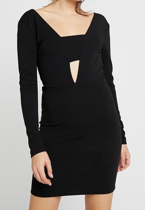 Kobieta ubrana w dopasowaną czarną sukienkę z długim rękawem, z geometrycznym wycięciem na klatce piersiowej i kwadratowym dekoltem.