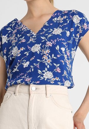 Blusa azul de cuello en V con estampado floral en blanco, rosa y naranja, llevada por dentro de pantalones de talle alto color marfil.
