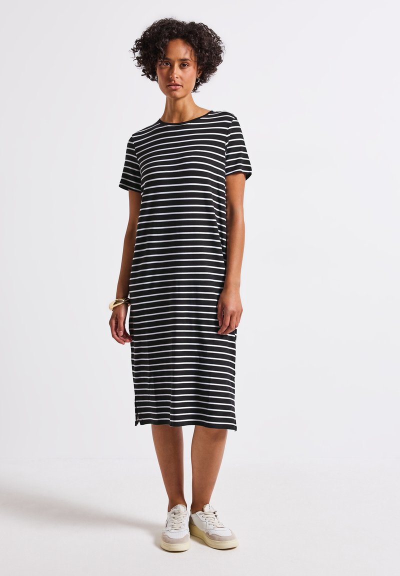Vestido midi a rayas en blanco y negro con mangas cortas, escote redondo y aberturas laterales. Lleva zapatillas blancas.