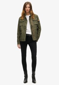 Olivengrøn arbejdsjakke med to brystlommer, knaplukning og en blød tekstur, lagdelt over en hvid skjorte og sorte stramme jeans.