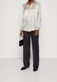 Camisa de botón de seda en dos tonos con cuello decorativo, combinada con pantalones de pierna ancha de encaje azul marino. Bolso negro, zapatos planos negros.