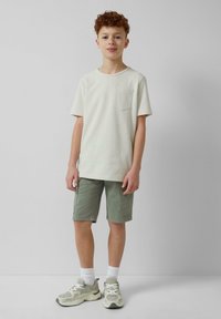 Lichtbeige T-shirt met korte mouwen en een borstzak, gecombineerd met olijfgroene shorts. Grijze sneakers met witte accenten en sportieve sokken.