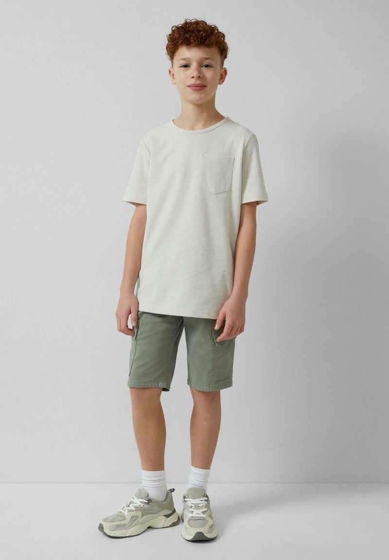 Lichtbeige T-shirt met korte mouwen en een borstzak, gecombineerd met olijfgroene shorts. Grijze sneakers met witte accenten en sportieve sokken.