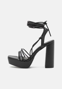 Las sandalias de plataforma negras cuentan con un tacón alto y grueso, punta abierta y un diseño de tiras entrelazadas con un envoltura de tobillo atada. Acabado suave y brillante.