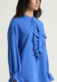 Blusa blu con scollo rotondo, dettaglio a volant cascante sul davanti e maniche lunghe con polsini arricciati. Tessuto dalla trama liscia.