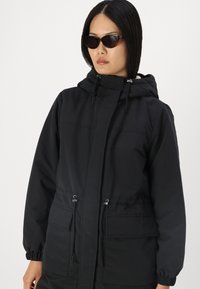 Veste noire imperméable avec capuche à cordon, poignets élastiques, deux poches avant et taille ajustable. Texture lisse avec un design minimaliste.