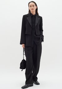 Blazer negro recortado con solapas de satén, combinado con una camisa a juego de botones y pantalones sueltos. Accesorios con un bolso negro texturizado.