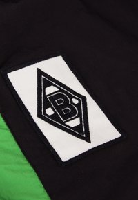 Zwarte stof met een witte rechthoekige patch waarop een zwart diamanten logo staat met een "B" in het midden, vergezeld van levendige groene accenten.