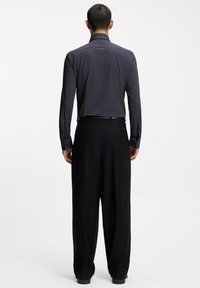 Uomo di spalle che indossa una camicia nera a maniche lunghe con pois bianchi e pantaloni neri larghi su uno sfondo semplice.