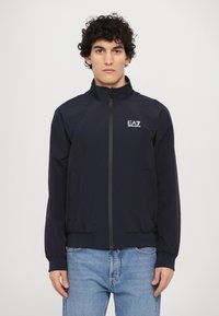 Chaqueta ligera de color azul oscuro con cuello alto, cierre de cremallera, bolsillos laterales y un pequeño logo blanco de EA7 en el lado izquierdo del pecho.