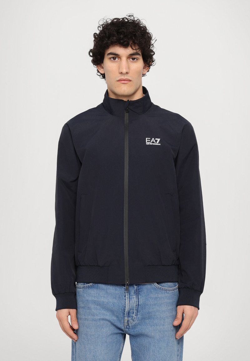 Chaqueta ligera de color azul oscuro con cuello alto, cierre de cremallera, bolsillos laterales y un pequeño logo blanco de EA7 en el lado izquierdo del pecho.