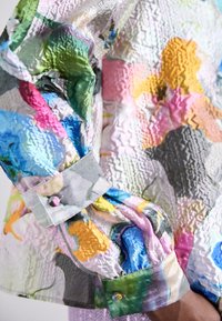 Multicolored blouse featuring a crinkled texture, vivid floral patterns in pink, blue, green, and yellow, with buttoned cuffs and a smooth finish.