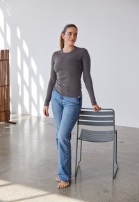 Graues Langarmshirt kombiniert mit blauer Jeans. Das Shirt hat einen figurbetonten Schnitt und eine glatte Textur. In der Nähe steht ein minimalistischer grauer Stuhl.