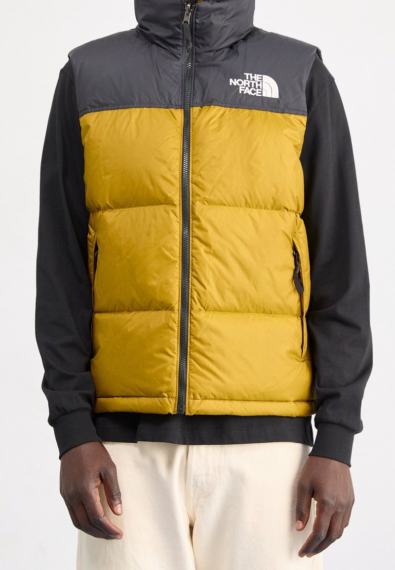 Yellow and gray insulated vest with a high collar, front zipper, two side pockets, and white logo on the chest. Black long-sleeve shirt underneath.