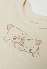 Svetle béžové tričko s potiskem dvou karikaturních štěňat se světle hnědými akcenty a textem "malí přátelé" pod ním.
