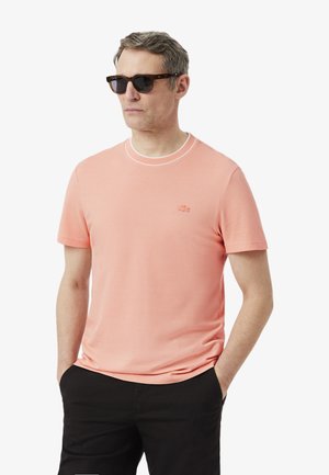 Hombre con gafas de sol oscuras, camiseta de manga corta color salmón con logo y pantalones negros, de pie con las manos en los bolsillos.
