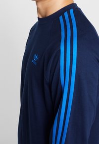 Camisa de manga larga color azul marino con tres franjas verticales color azul claro en cada hombro y un pequeño logo azul en relieve en el pecho.