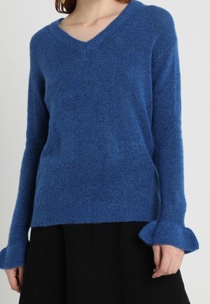 Femme portant un pull bleu à col en V avec des poignets à volants et une jupe noire sur un fond clair uni.