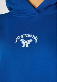 Modra kapuca iz bombaža, ki ima na prsih bel grafični logotip in dizajn metulja. Mehka tekstura, z vrvico za nastavitev kapuce.