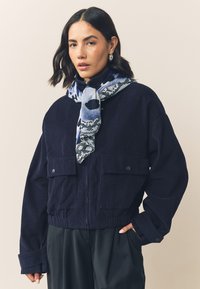 Navy corduroy cropped jacket with two front pockets, elastic waistband, and a blue and white patterned scarf around the neck.