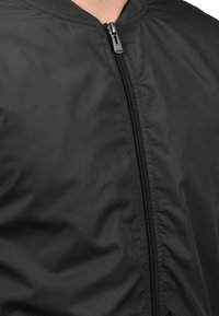 Mustakainen pommikaukkupaita, joka on valmistettu sileästä ja kevyestä materiaalista. Rintatasku on täysin vetoketjullinen ja kauluksessa on pyöreä muoto.