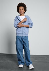 Sweatshirt azul-claro com fecho à frente e um bolso frontal, combinado com jeans azuis de corte largo e tênis brancos. T-shirt branca simples por baixo.