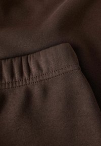 Brunt stoff med en glatt tekstur, med elastisk midjebånd og synlig søm-detalj. Materialet virker mykt og elastisk.