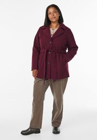 Burgundy belted coat with large collar and button closure, paired with brown pants and black ankle boots, featuring a smooth texture.