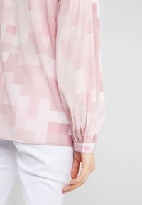 Blouse à manches longues à carreaux rose clair avec poignets boutonnés portée avec un pantalon blanc, montrant le côté et l'avant-bras d'une personne.