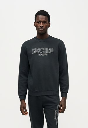 Мъж представя черен суичър Moschino Active и подходящо долнище на анцуг на изчистен светъл фон.