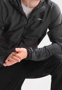 Svart lättviktsjacka med huva, dragkedjestängning och elastiska ärmslut. Har en grå logotyp på bröstet och en slät, matt tygtextur.